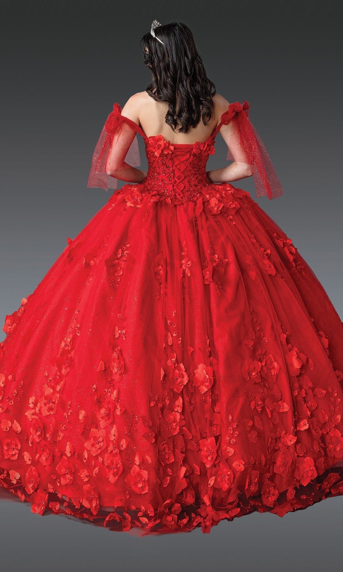 A person wearing the Quinceanera Dress 1792 By Dancing Queen—a stunning red gown with beaded floral appliques, off-the-shoulder sleeves, and a tiara—is shown from the back against a gray background.